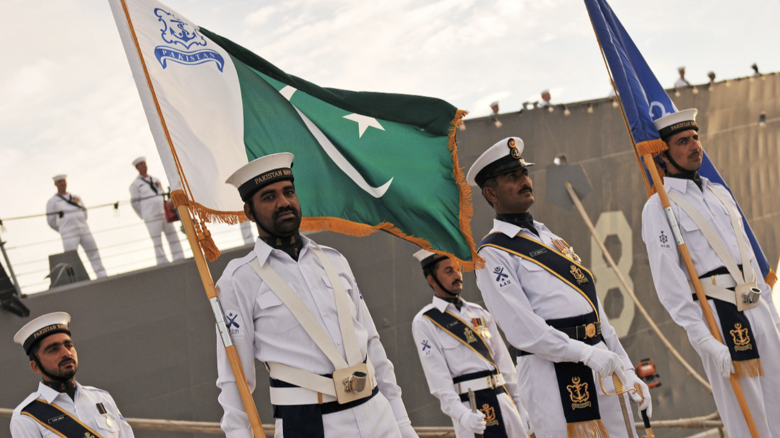 Pakistani sailors stand at attention in front of a ship. One sailor is holding the flag of Pakistan.