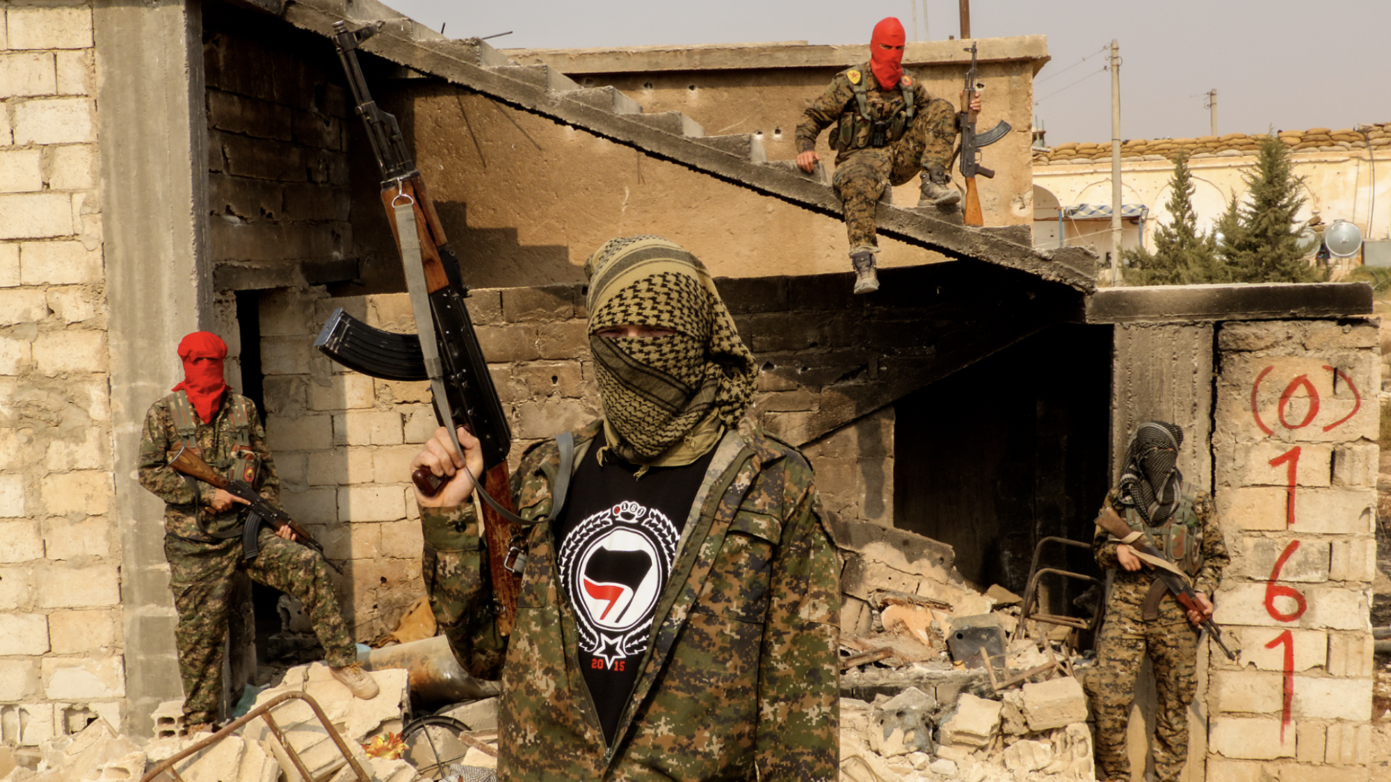 Four men in camouflage clothing and scarves around their heads stand in front of rubble holding guns.