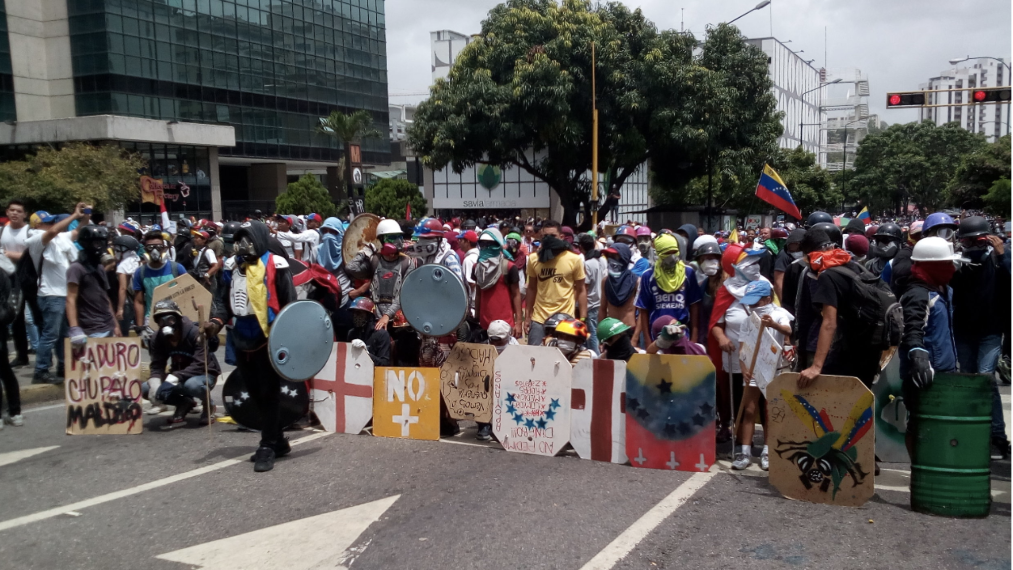 A crowd stands in the middle of a street wearing gas masks and holding signs with the Venezuelan flag.