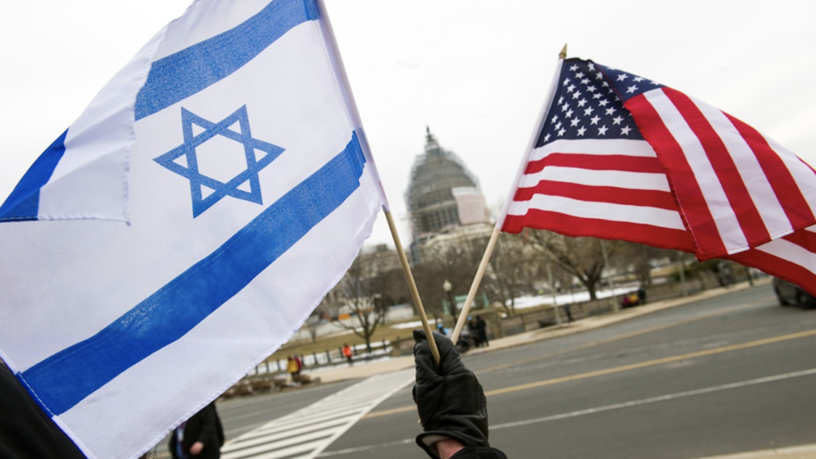 A hand waving the flags of the U.S. and Israel in front of the U.S. capitol building