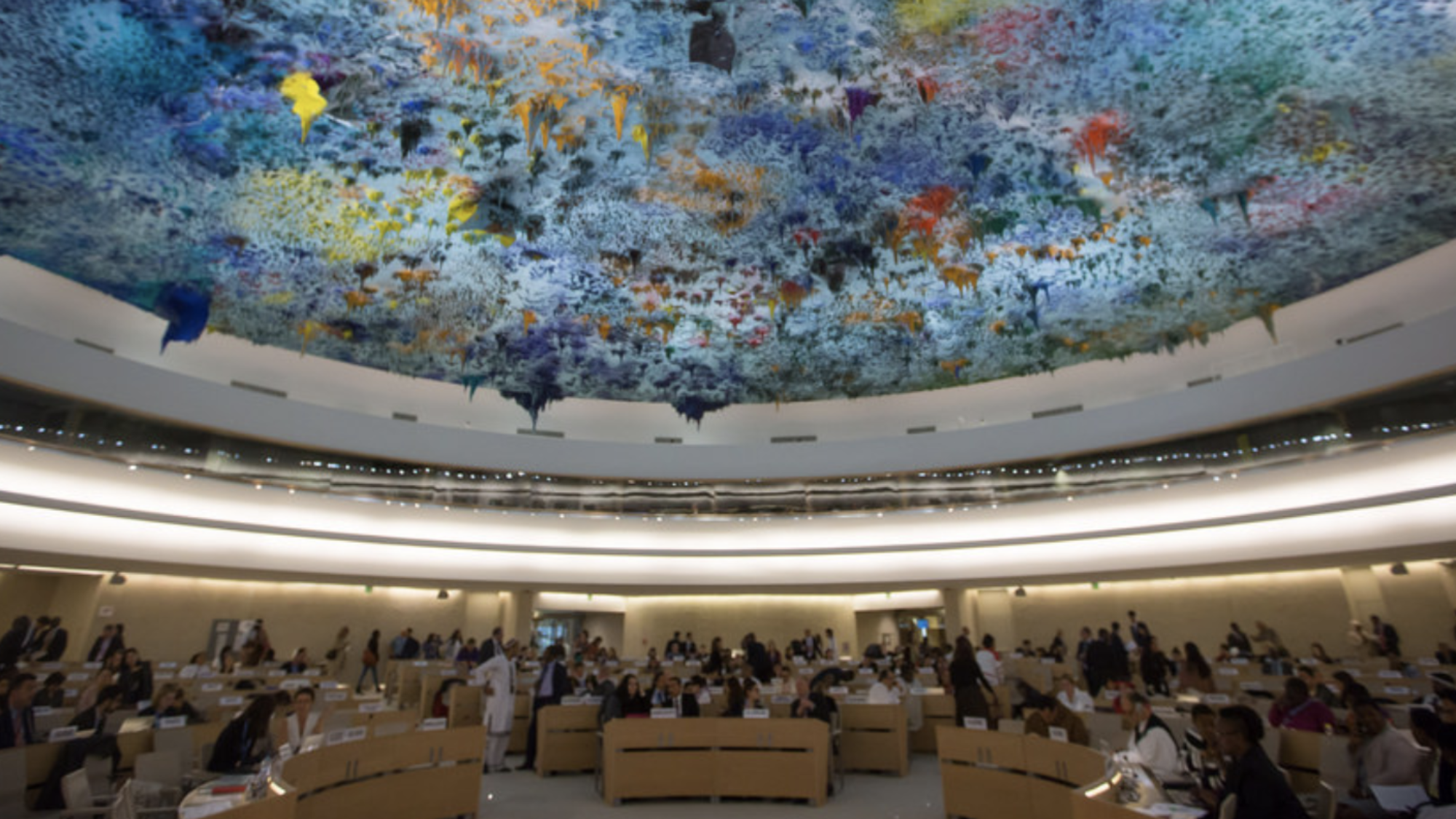 Lawmakers sit at tables in a large circular room with a colorful ceiling