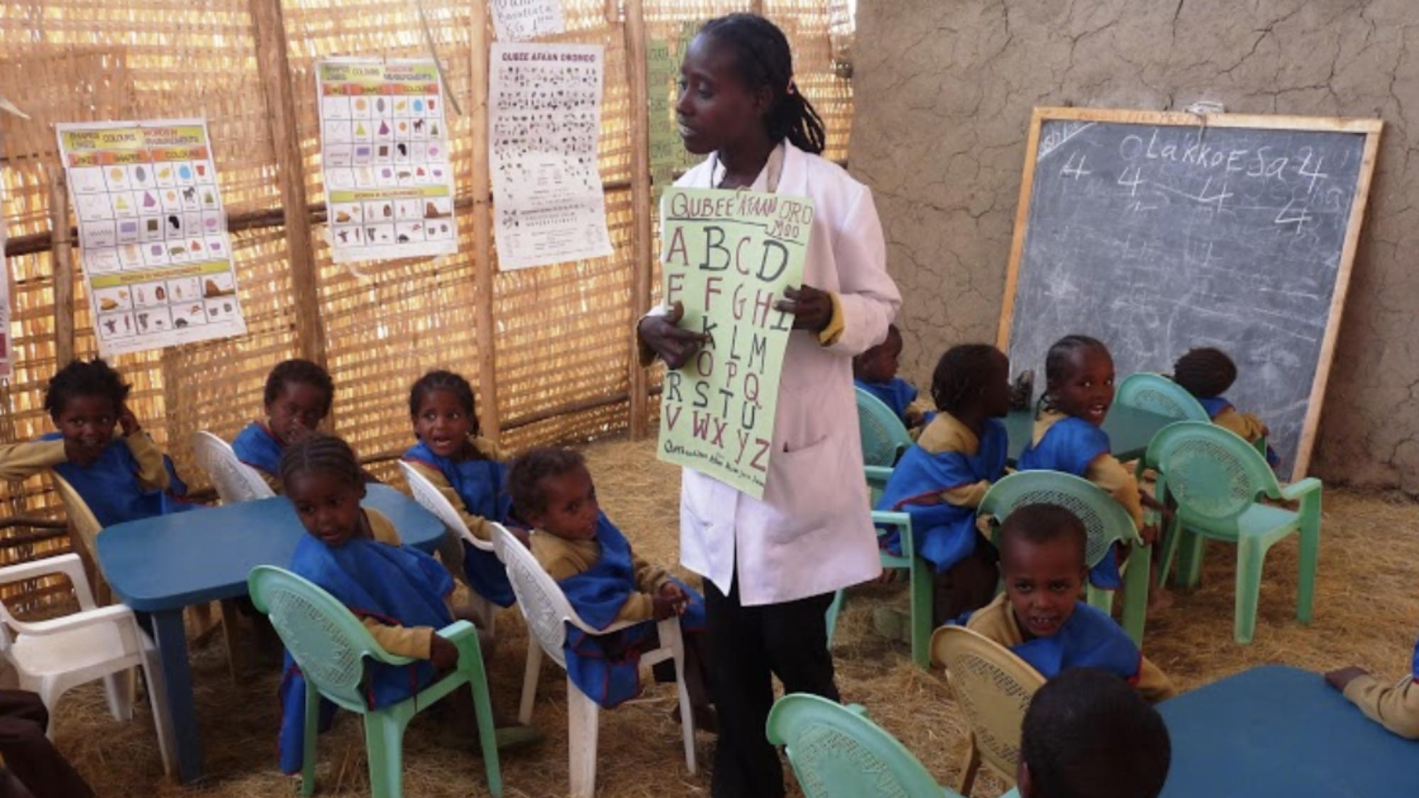 A teacher shows a poster with the English alphabet to a classroom of young students.