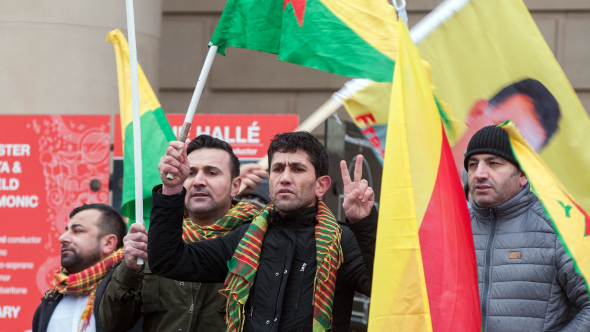 Four men holding Kurdish flags, participating in protest against Kurdish military aggression.