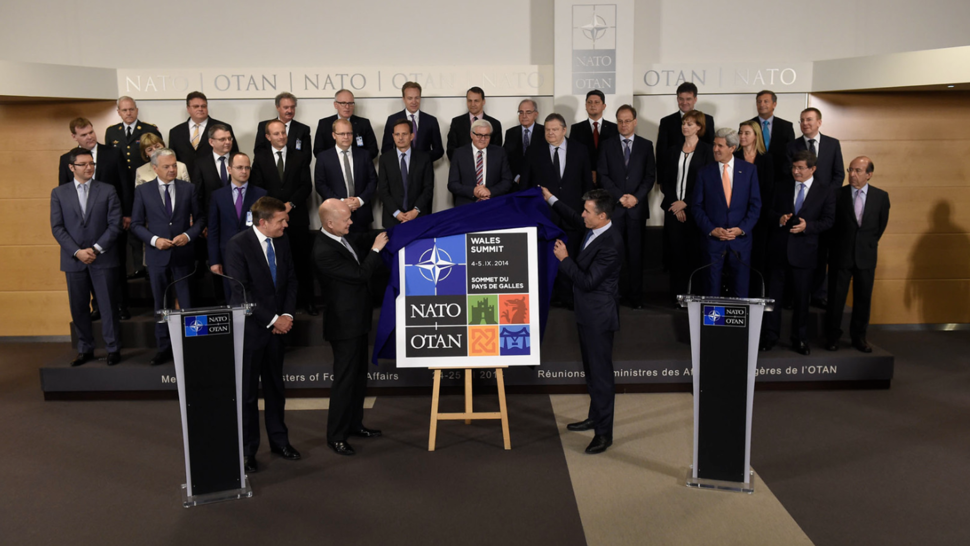 Leaders of NATO countries stand on a stage and look on as the Secretaries of State from the UK and Wales unveil the logo of the 2014 NATO Summit in Wales.