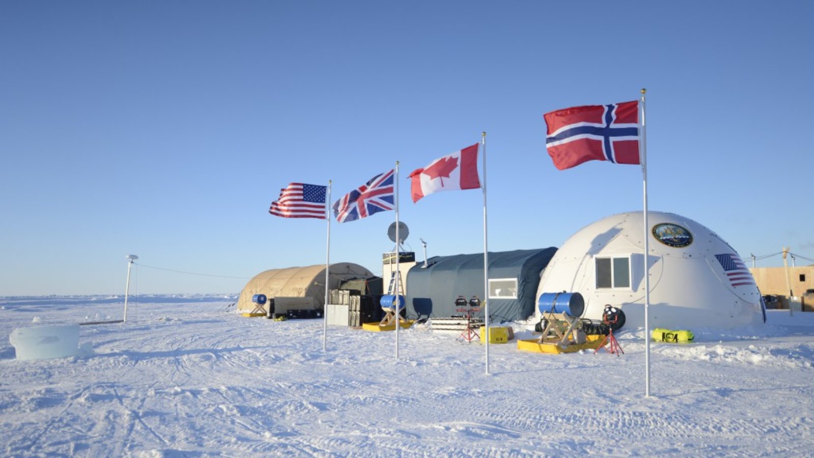 The flags of the U.S., the UK, Norway, and Canada are planted next to each other in the ice outside of a research station in the Arctic.