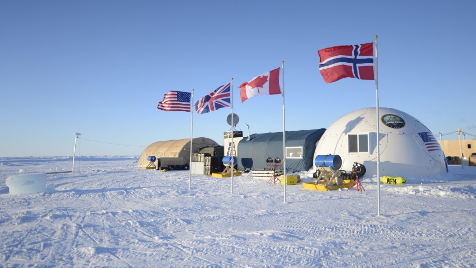 The flags of the U.S., the UK, Norway, and Canada are planted next to each other in the ice outside of a research station in the Arctic.