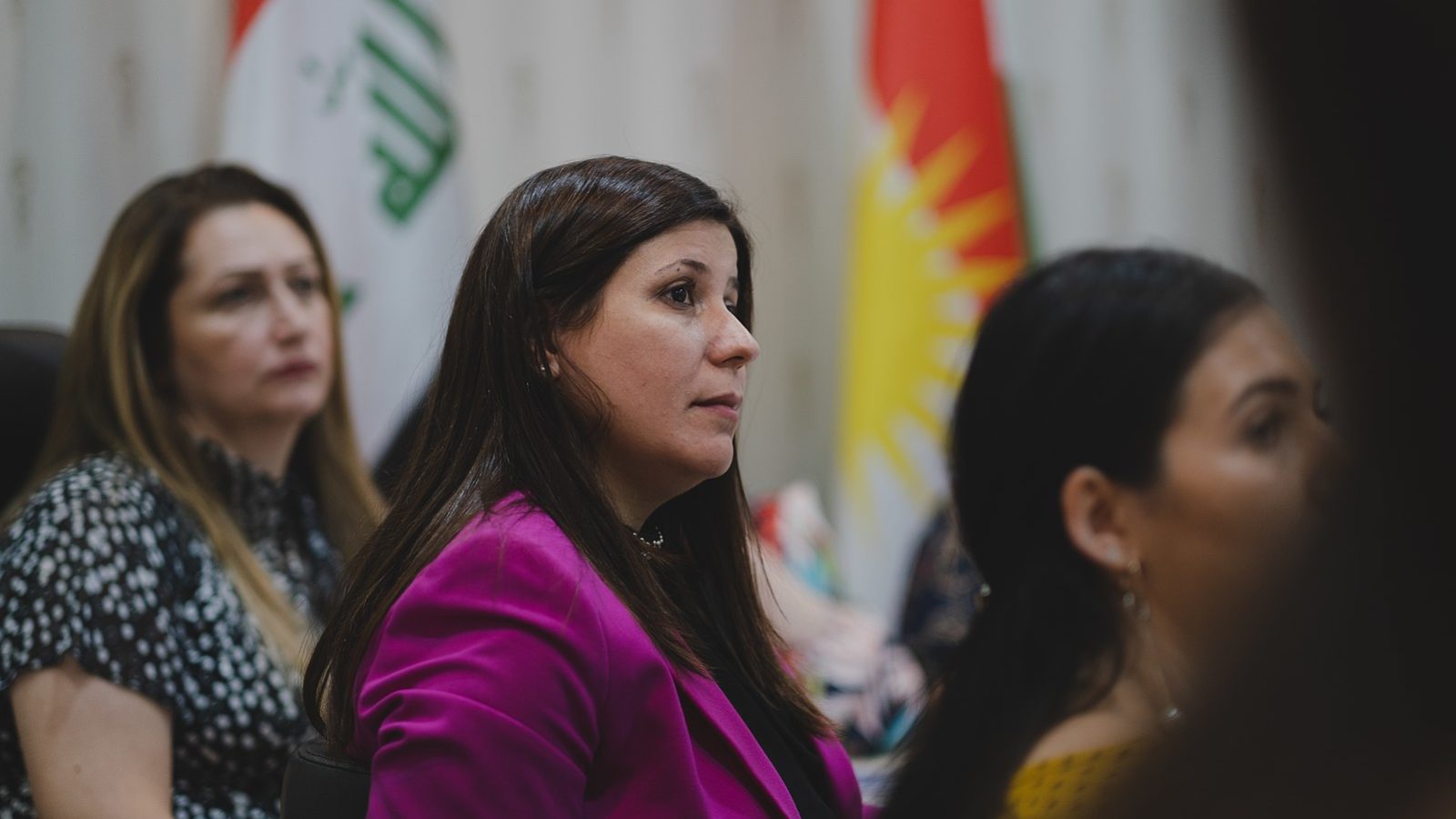 Florin Gorgis, a female human rights and women&#039;s rights activist, sits at a table looking into the distance. The flags of Kurdistan and Iraq are in the background.