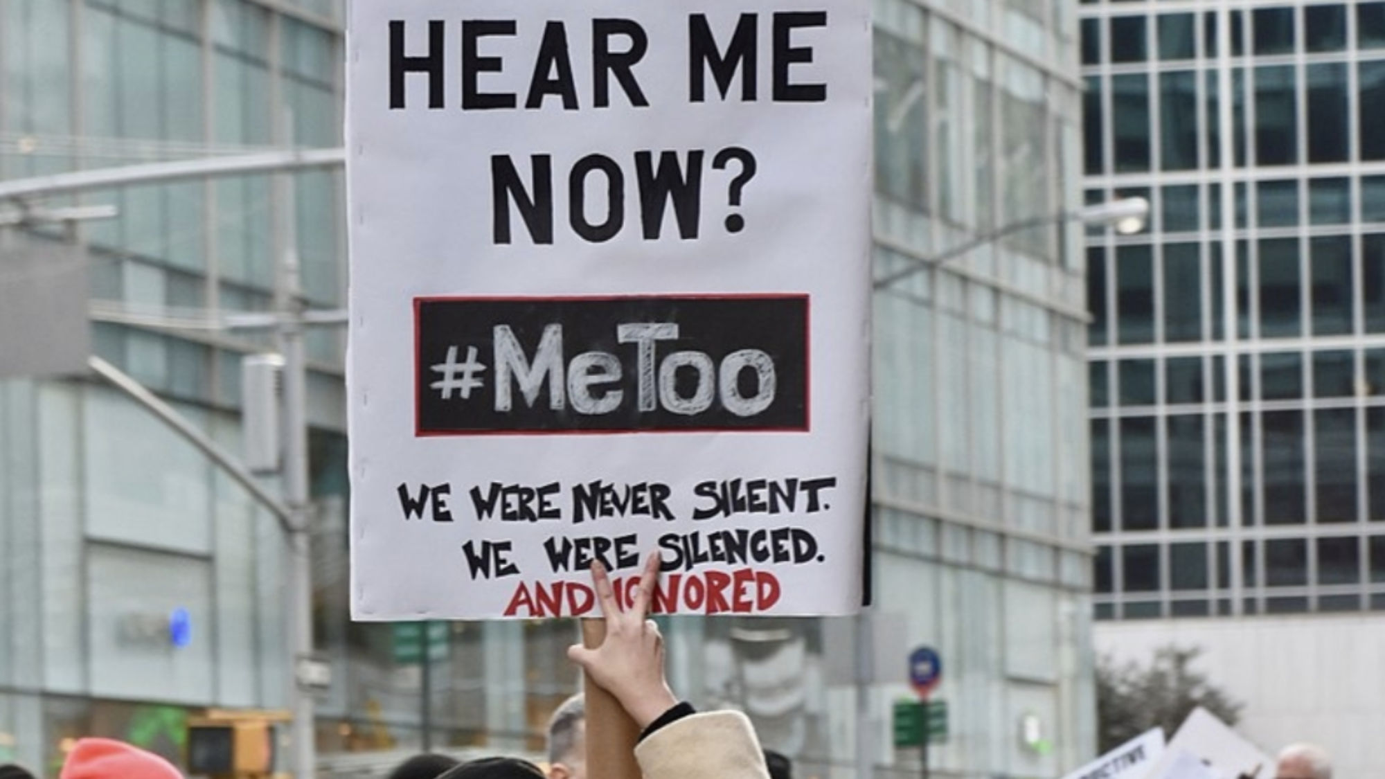 A woman stands in a crowd of protestors holding a sign that reads, &quot;Can you hear me now? #MeToo. We were never silent. We were silenced. And ignored.&quot;