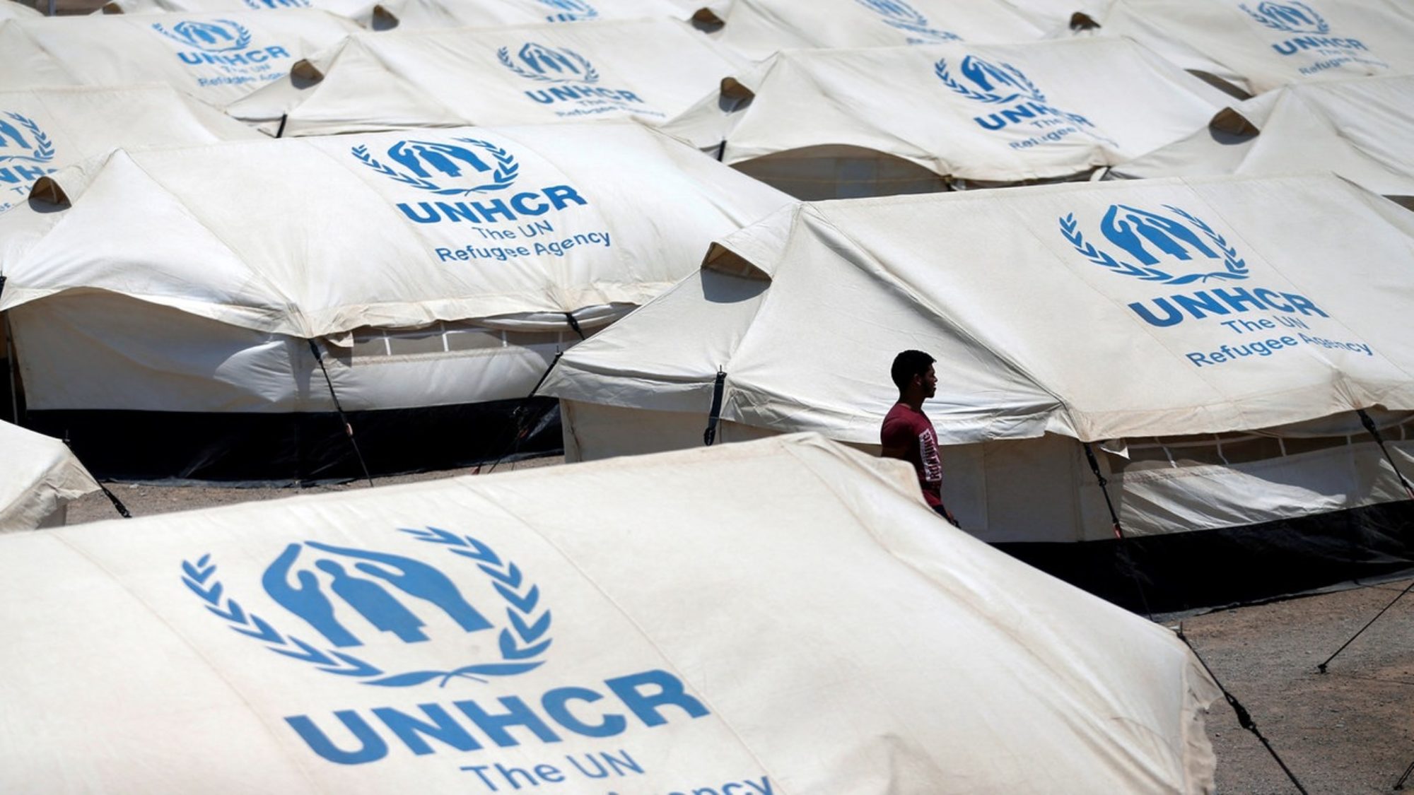 A man walks alone through a cluster of white tents with the UNHCR logo on them