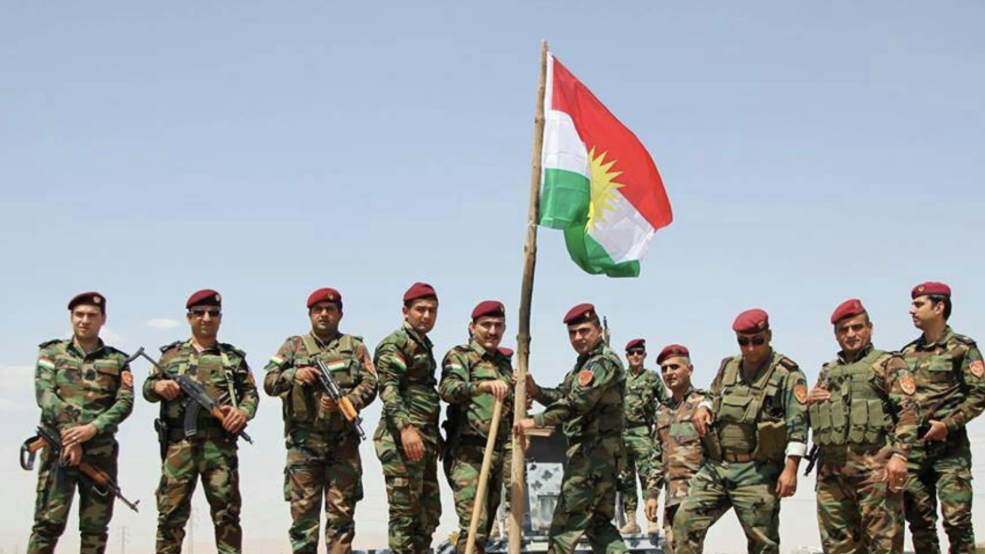 A group of soldiers stand on rocky soil holding the Kurdish flag