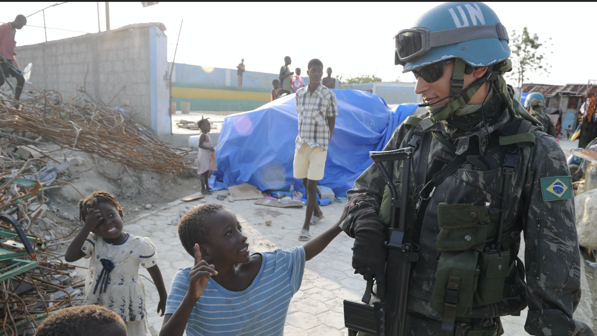 A tall male wearing a UN peacekeeper helmet says hello to some small children in Brazil.