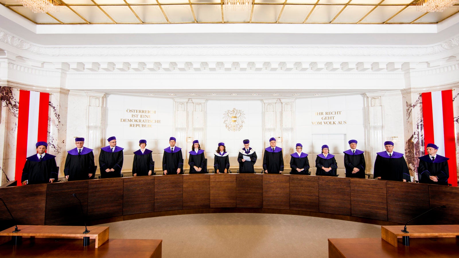 Members of the Austrian constitutional court stand behind a long wooden table flanked by the Austrian flag.