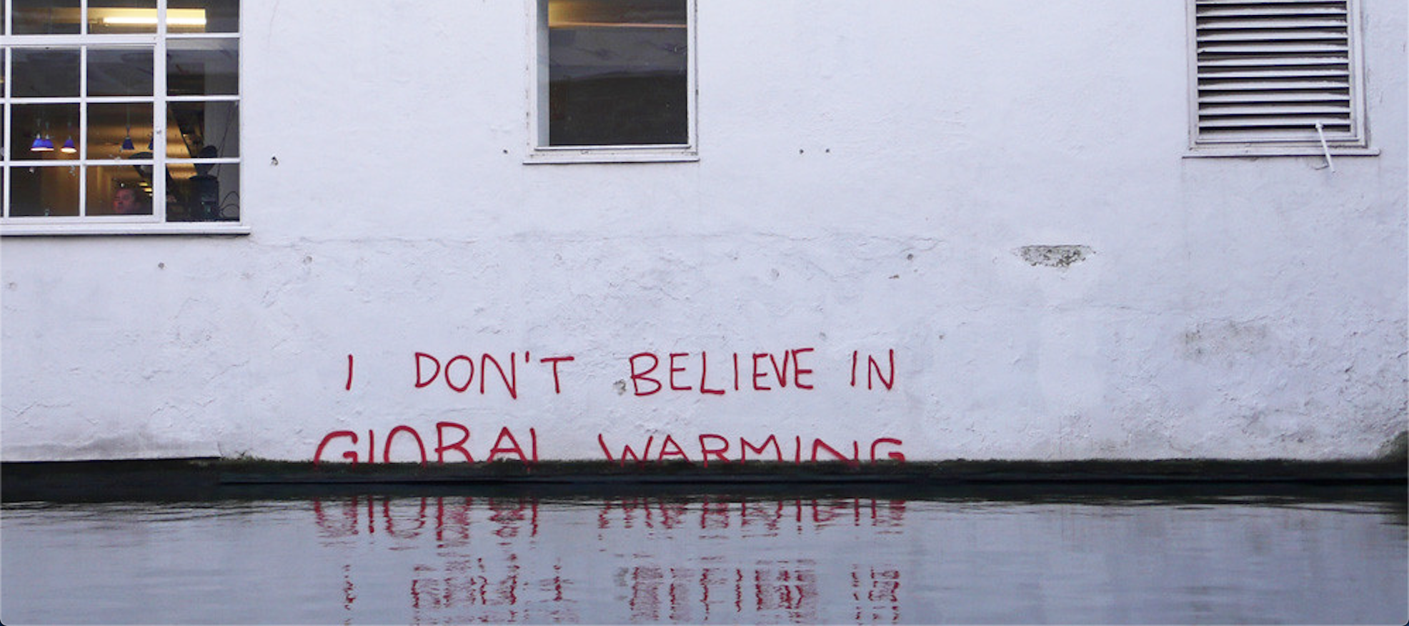 Red graffiti reads &quot;I don&#039;t believe in global warming&quot; on a wall that is submerged in water