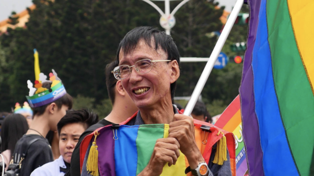 Activist Chi Chia-wei smiles holding a rainbow striped flag with a rainbow striped shirt.