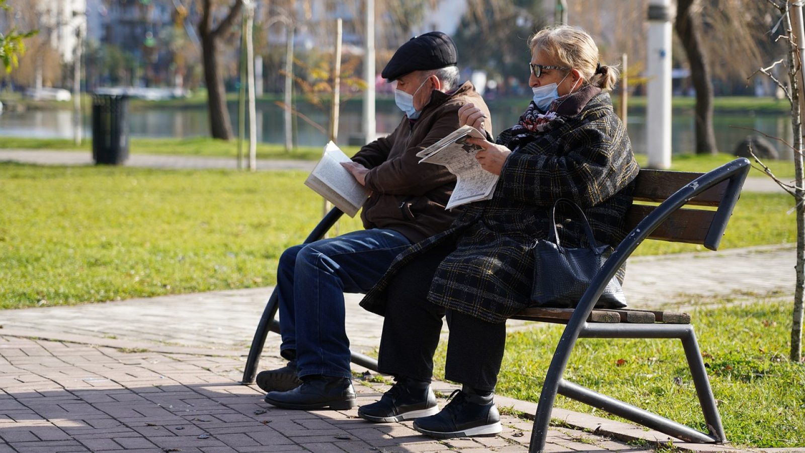 two elderly people with masks on sit on a bench outside