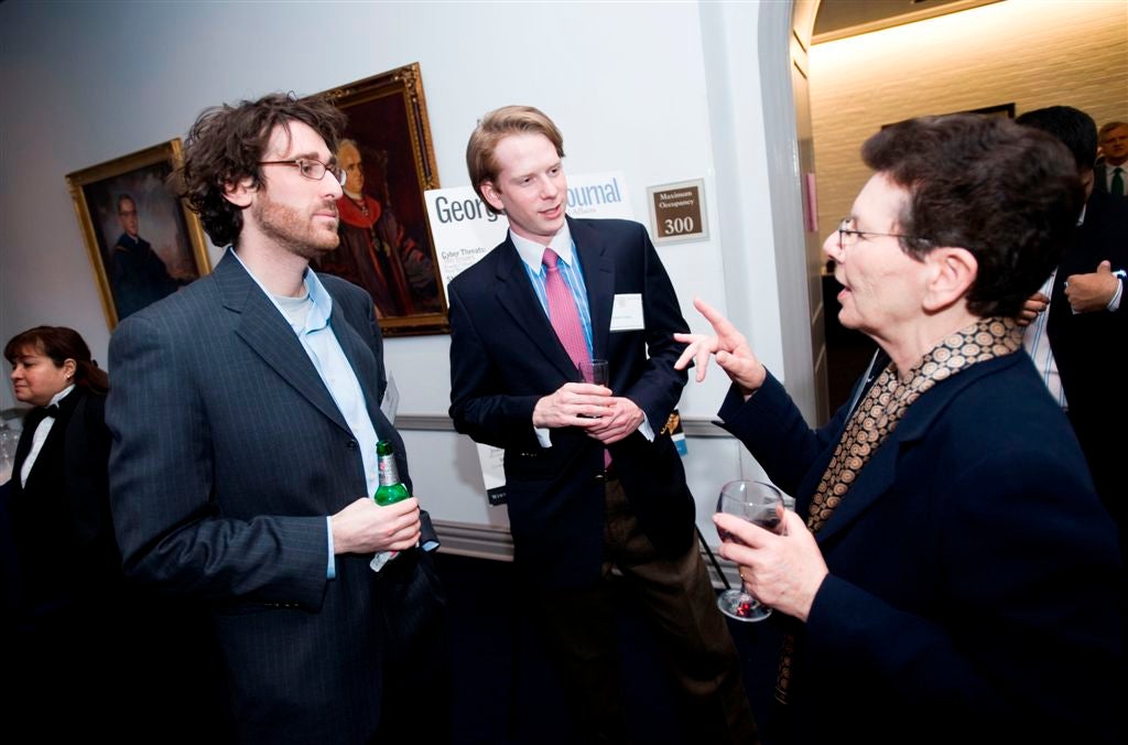 Three people in conversation at an event, one gesturing with a glass in hand, with framed paintings on the wall.