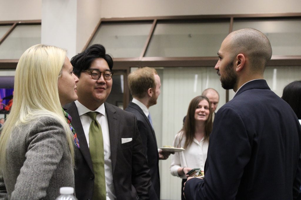 Three people chatting at a formal event, dressed in business attire, with others mingling in the background.