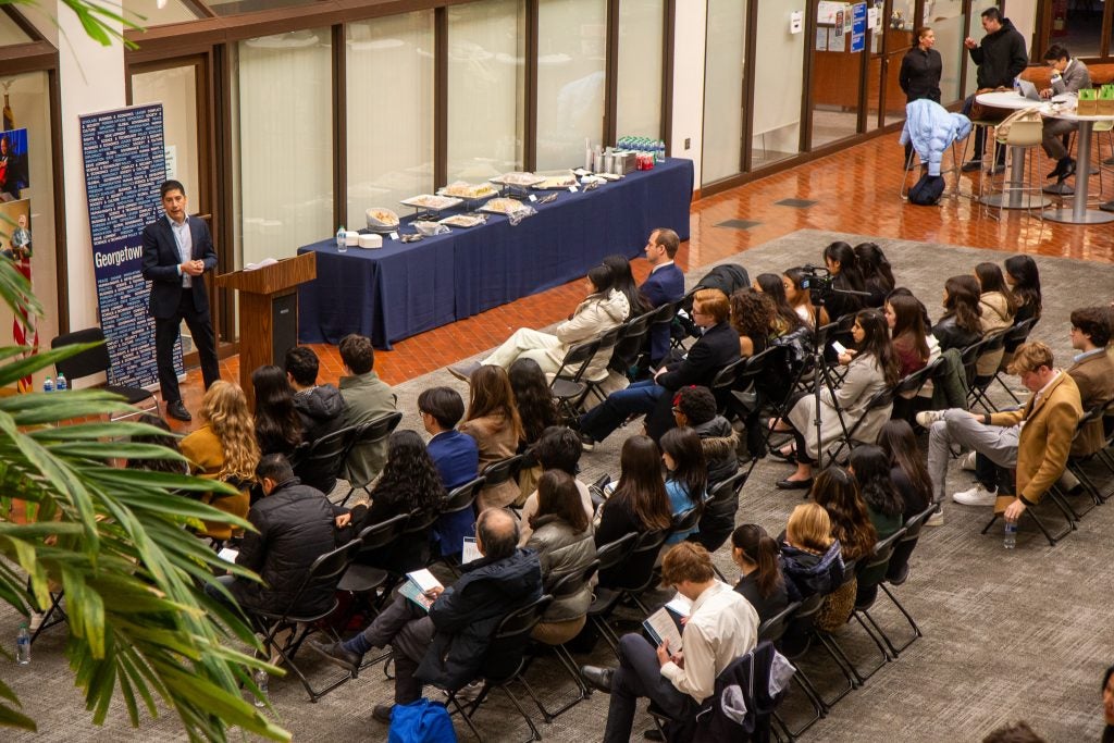 An overhead view of an event where a speaker addresses an audience seated in rows. A table with refreshments is set up nearby.