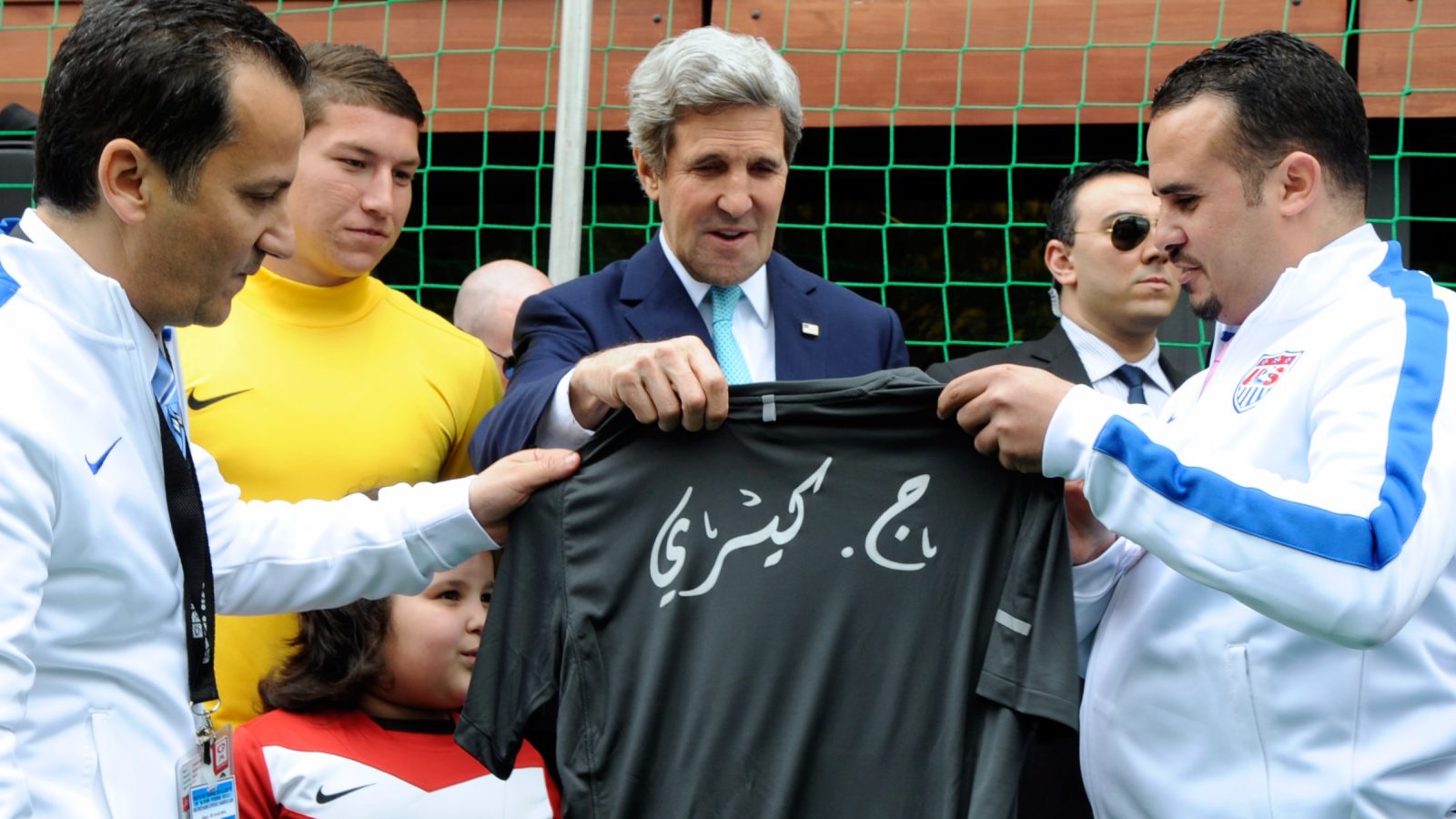 Three men holding a tshirt, one of them is a US Sec of State surrounded by sports executives from Algiers