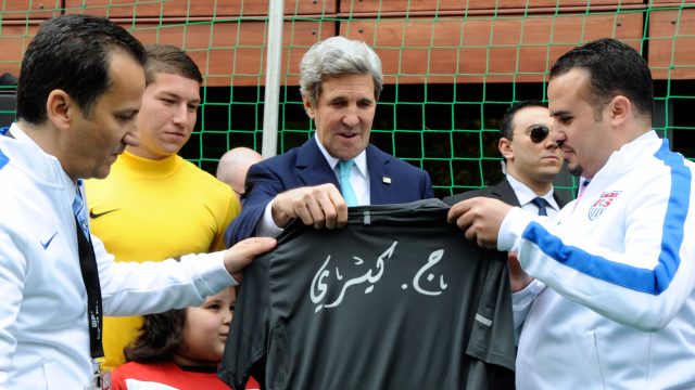 Three men holding a tshirt, one of them is a US Sec of State surrounded by sports executives from Algiers