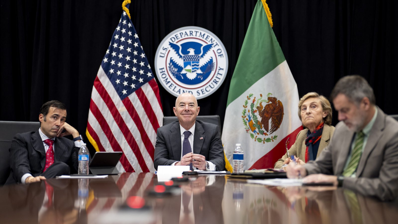 Homeland Secretary seated on a table with Mexico&#039;s Secretary of National Defense, General Luis Cresencio Sandoval, and the US Ambassador to Mexico, Ken Salazar. US and Mexico flags can be seen in the background.