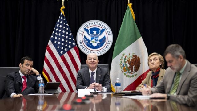 Homeland Secretary seated on a table with Mexico&#039;s Secretary of National Defense, General Luis Cresencio Sandoval, and the US Ambassador to Mexico, Ken Salazar. US and Mexico flags can be seen in the background.