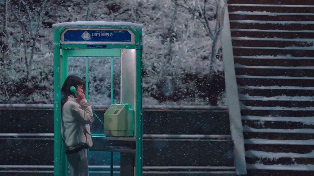 A person stands inside a green phone booth, holding a green telephone receiver to their ear, on a snowy night. The phone booth has a sign with Korean text at the top. Snow is falling gently, covering the ground, trees, and nearby staircase in a light blanket of white. The scene appears calm and serene, with the person seemingly engaged in a conversation amidst the quiet snowfall.