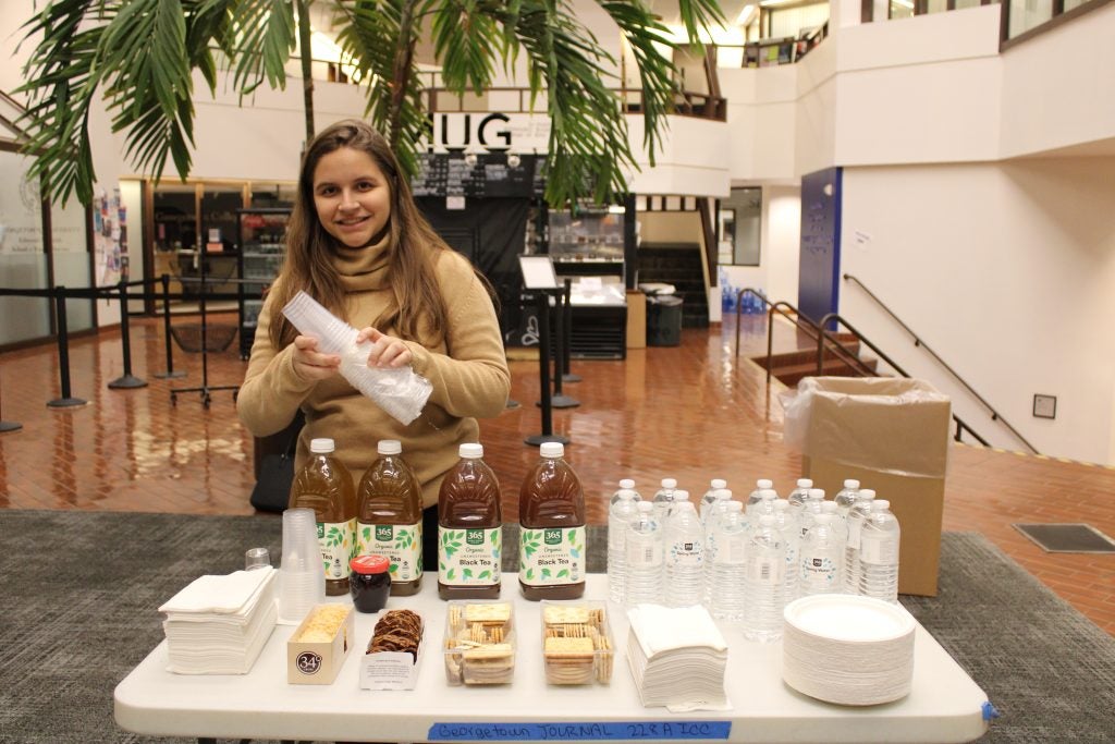 Woman smiling behind a table with drinks and snacks in a large indoor space with plants and seating in the background.