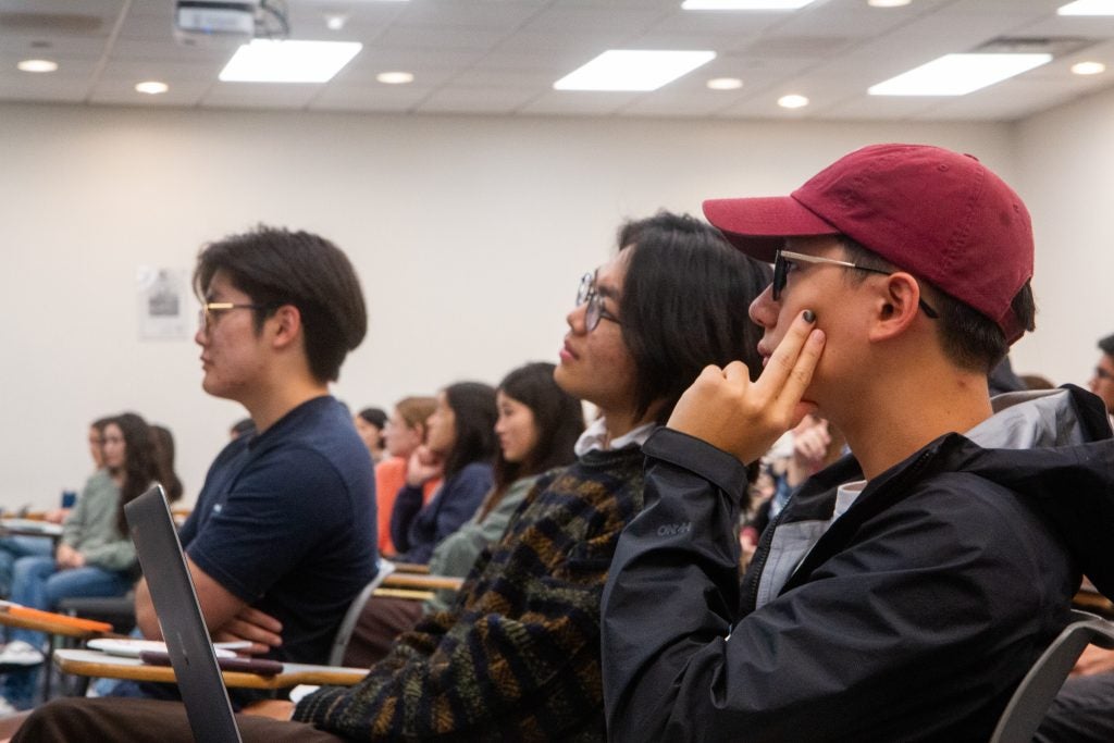 Students in a classroom listen attentively, with some taking notes. A student in a red cap is focused, resting his face on his hand.