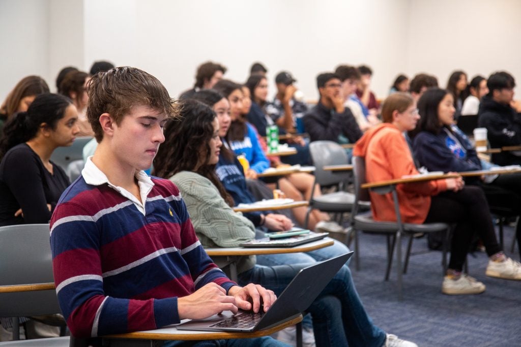 A classroom with students seated at desks, focused on a lecture. One in the foreground types on a laptop.