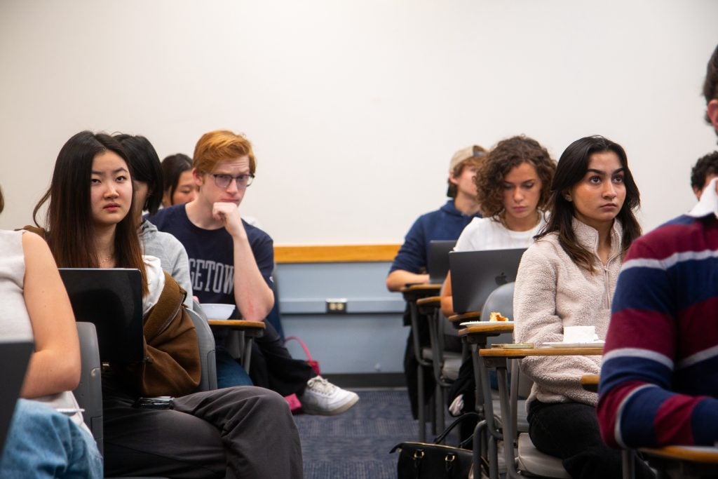 Students in a classroom, attentively listening, with some taking notes or using laptops.