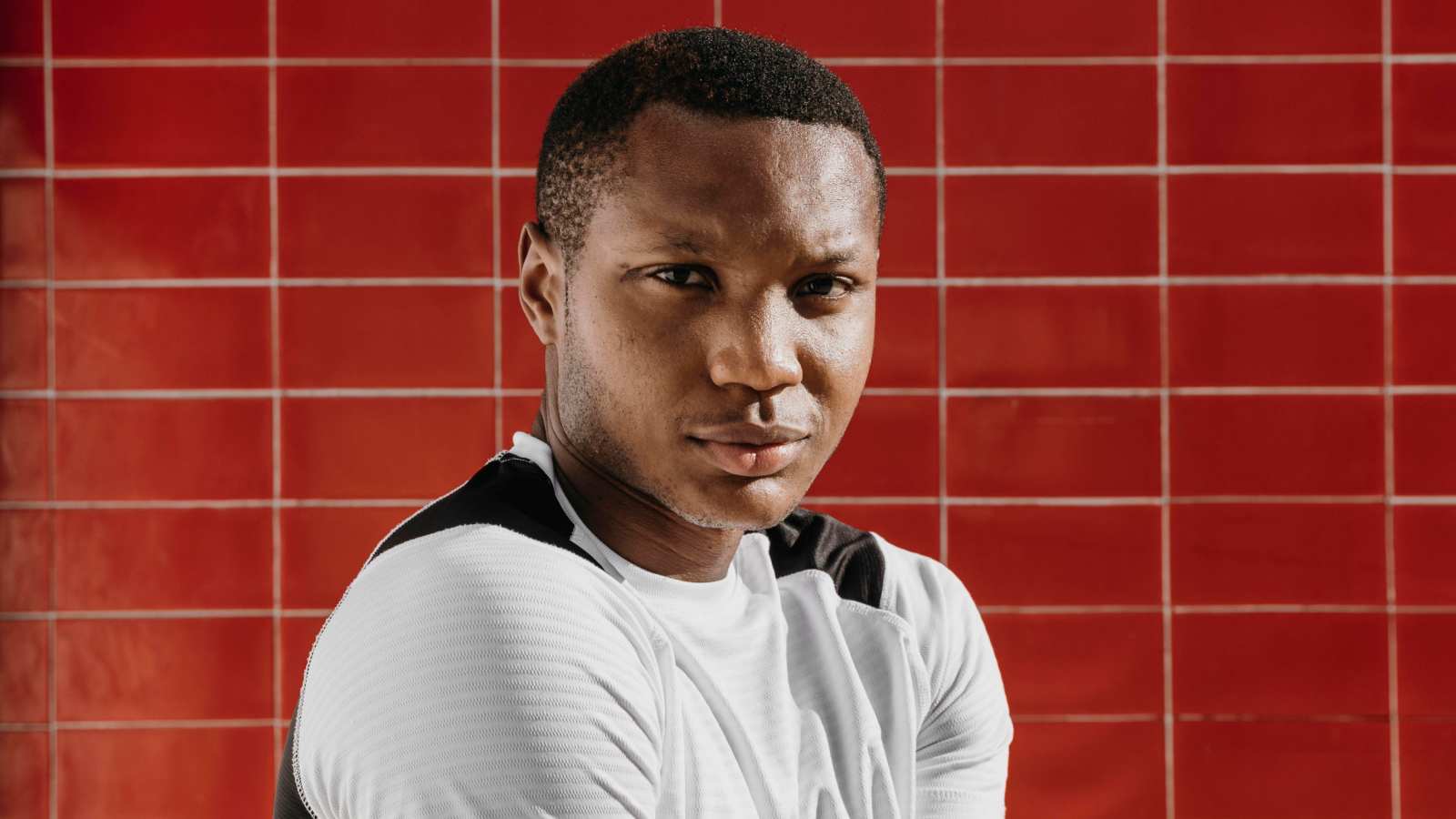 This image features a young man standing in front of a red tiled wall. He is wearing a white sports shirt with black stripes, looking directly at the camera with a subtle, serious expression.