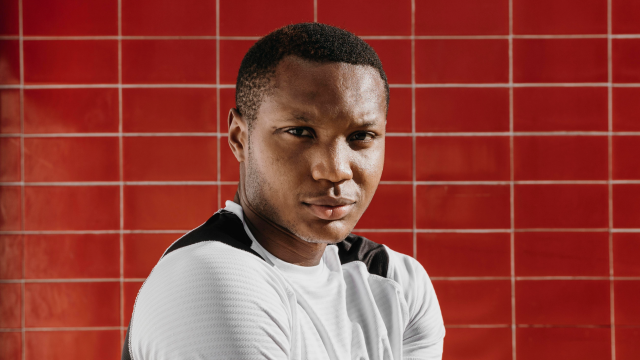 This image features a young man standing in front of a red tiled wall. He is wearing a white sports shirt with black stripes, looking directly at the camera with a subtle, serious expression.