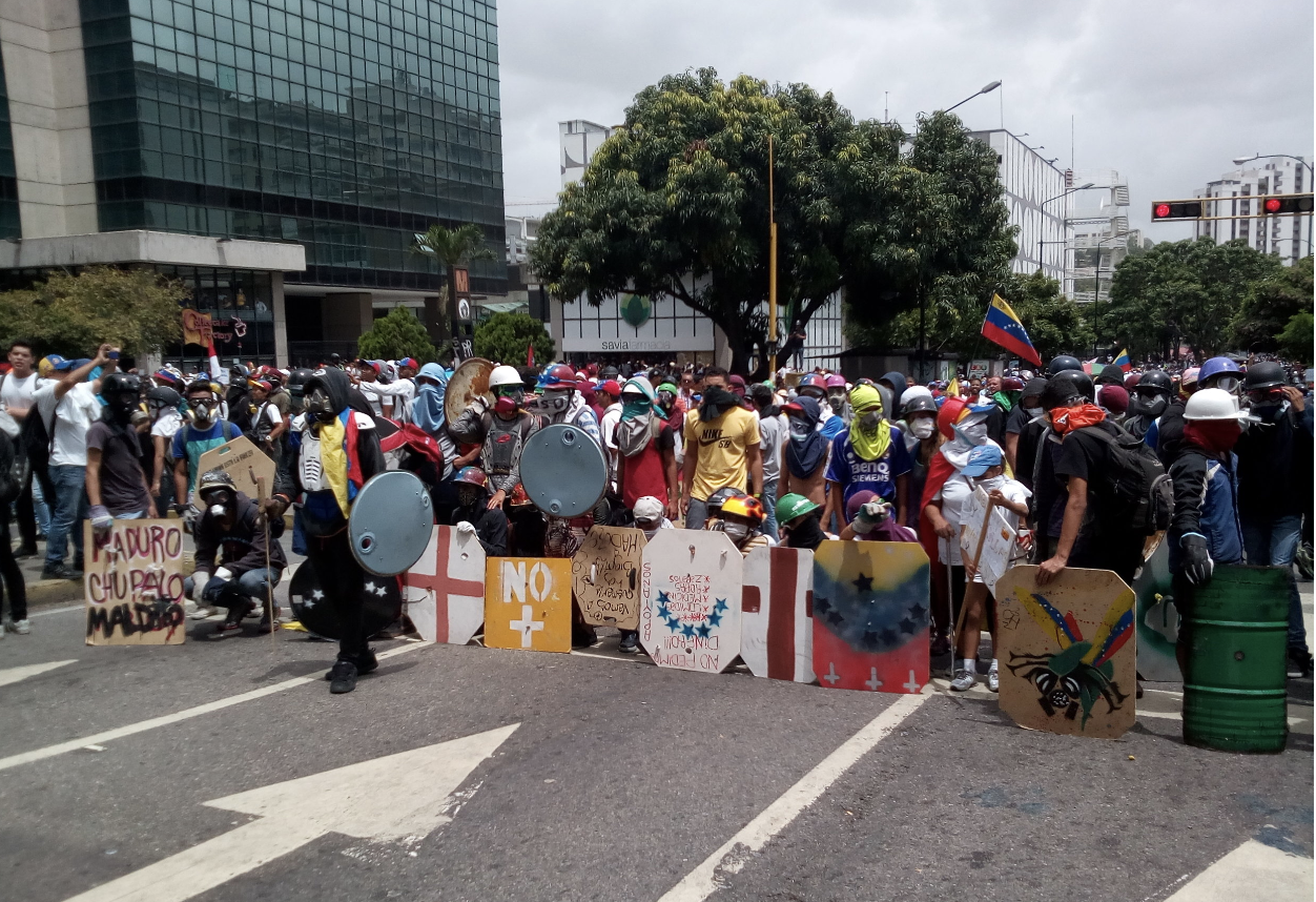 A crowd stands in the middle of a street wearing gas masks and holding signs with the Venezuelan flag.
