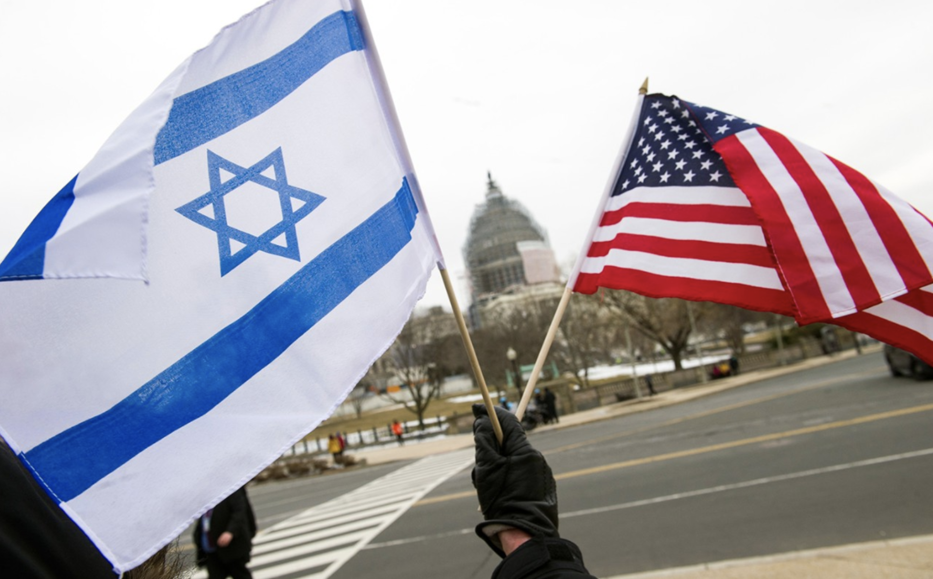 A hand waving the flags of the U.S. and Israel in front of the U.S. capitol building