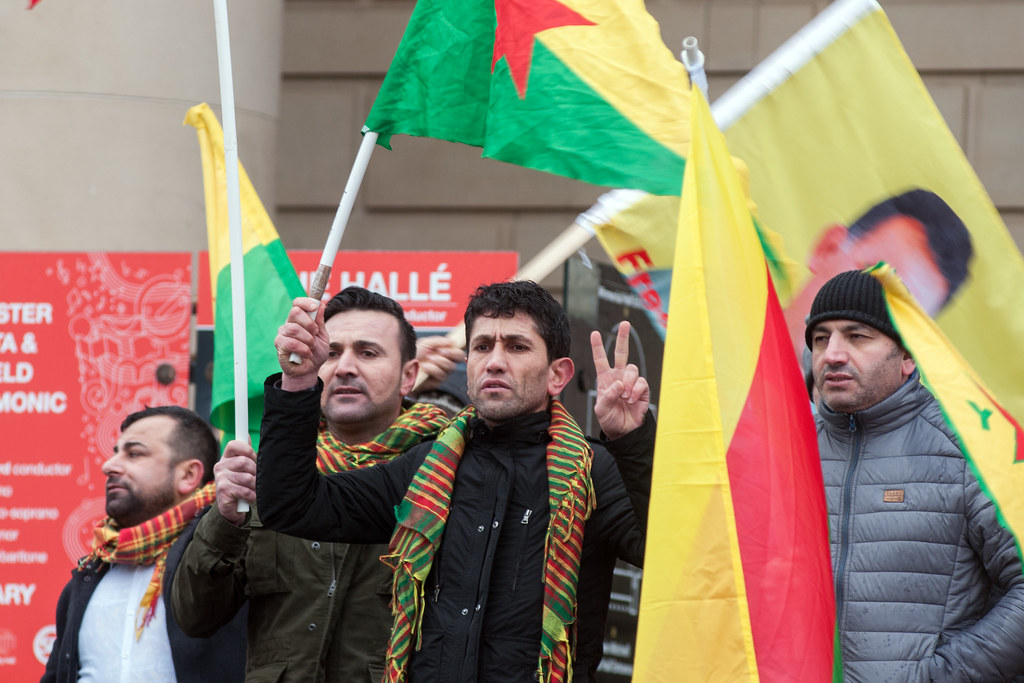 Four men holding Kurdish flags, participating in protest against Kurdish military aggression.