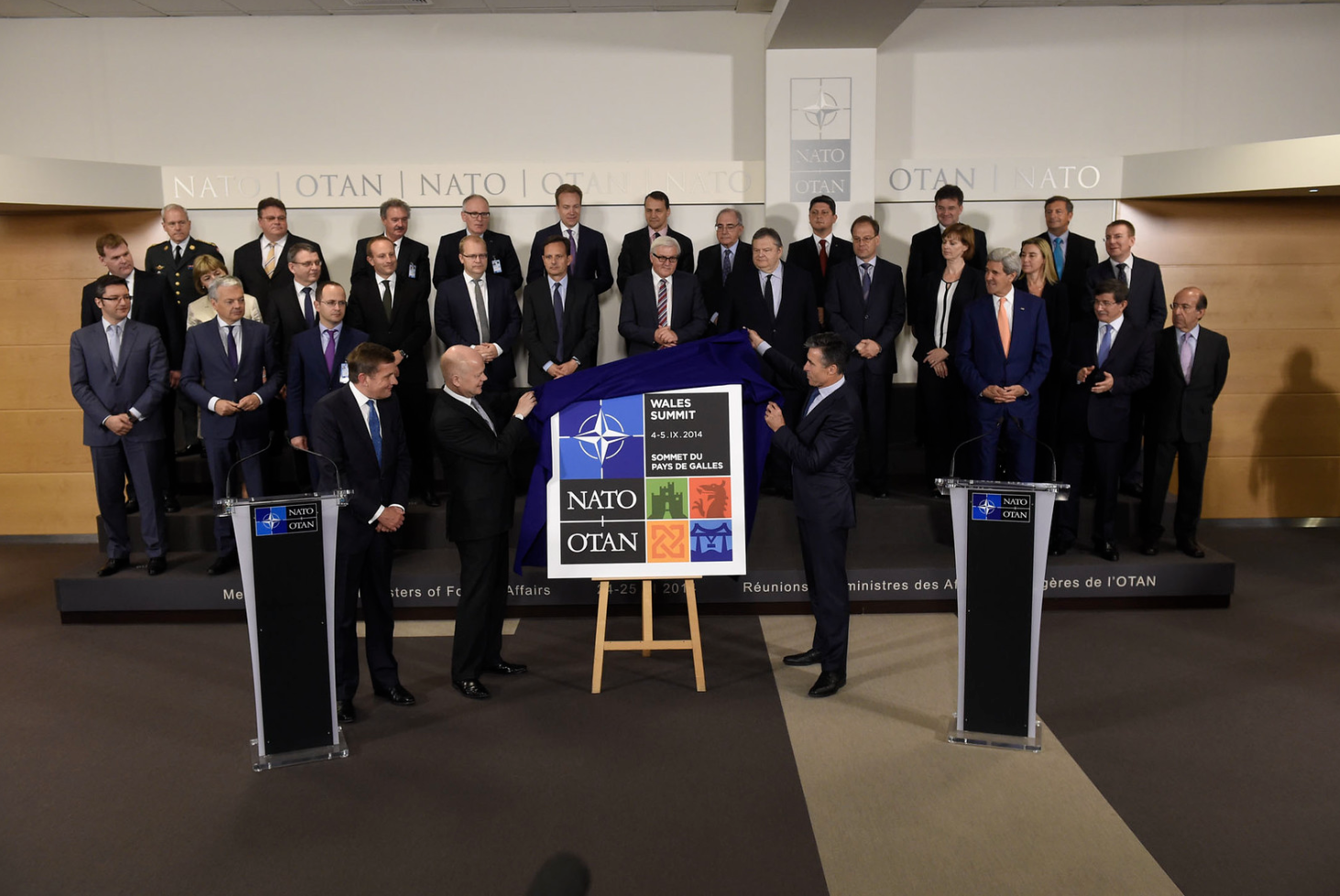 Leaders of NATO countries stand on a stage and look on as the Secretaries of State from the UK and Wales unveil the logo of the 2014 NATO Summit in Wales.