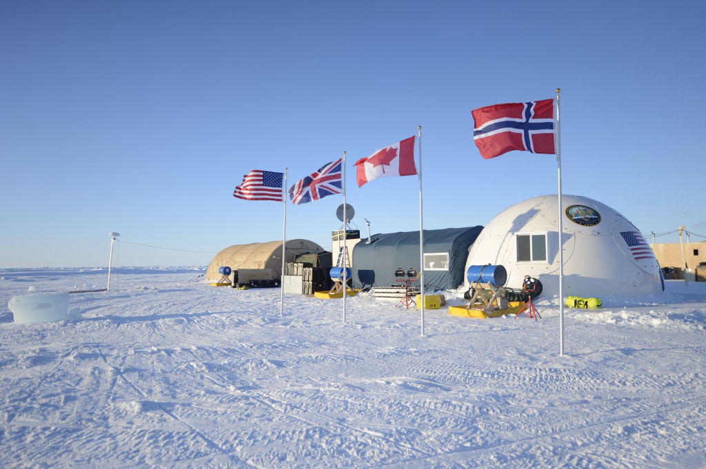 The flags of the U.S., the UK, Norway, and Canada are planted next to each other in the ice outside of a research station in the Arctic.