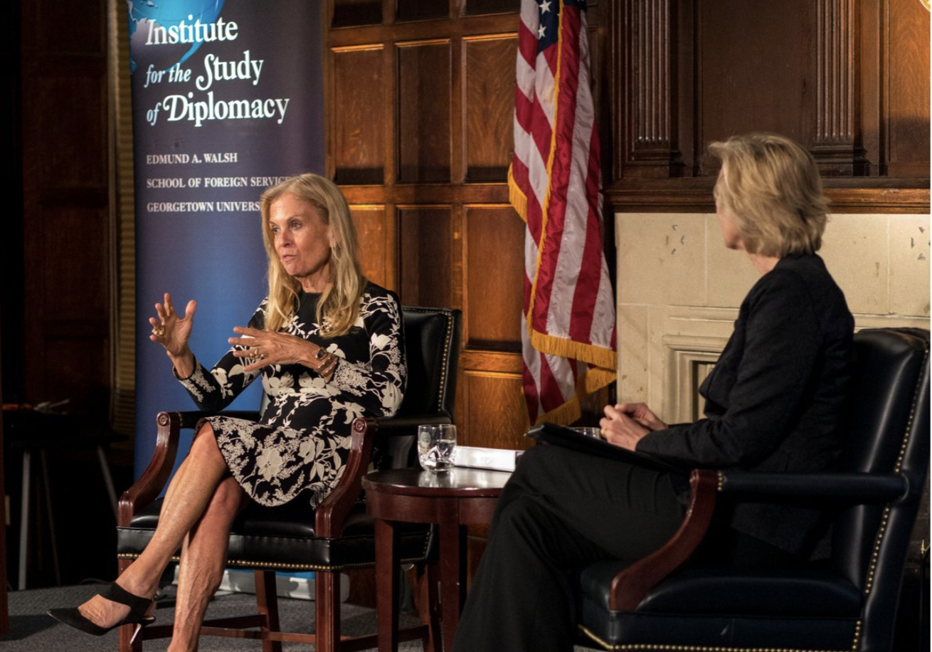 French Ambassador Jane Hartley sits on a stage in front of an American flag and a sign that reads, 
