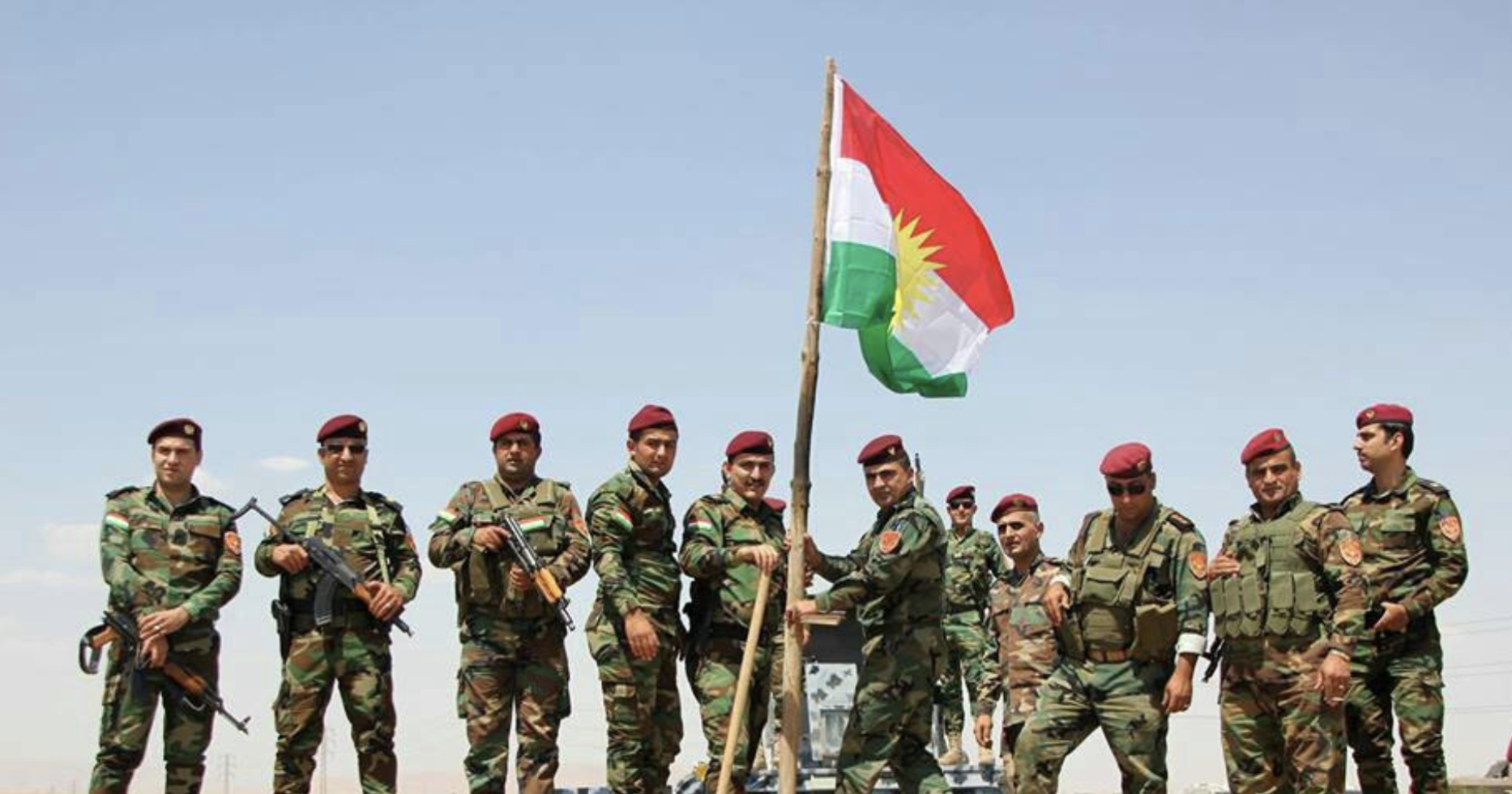 A group of soldiers stand on rocky soil holding the Kurdish flag