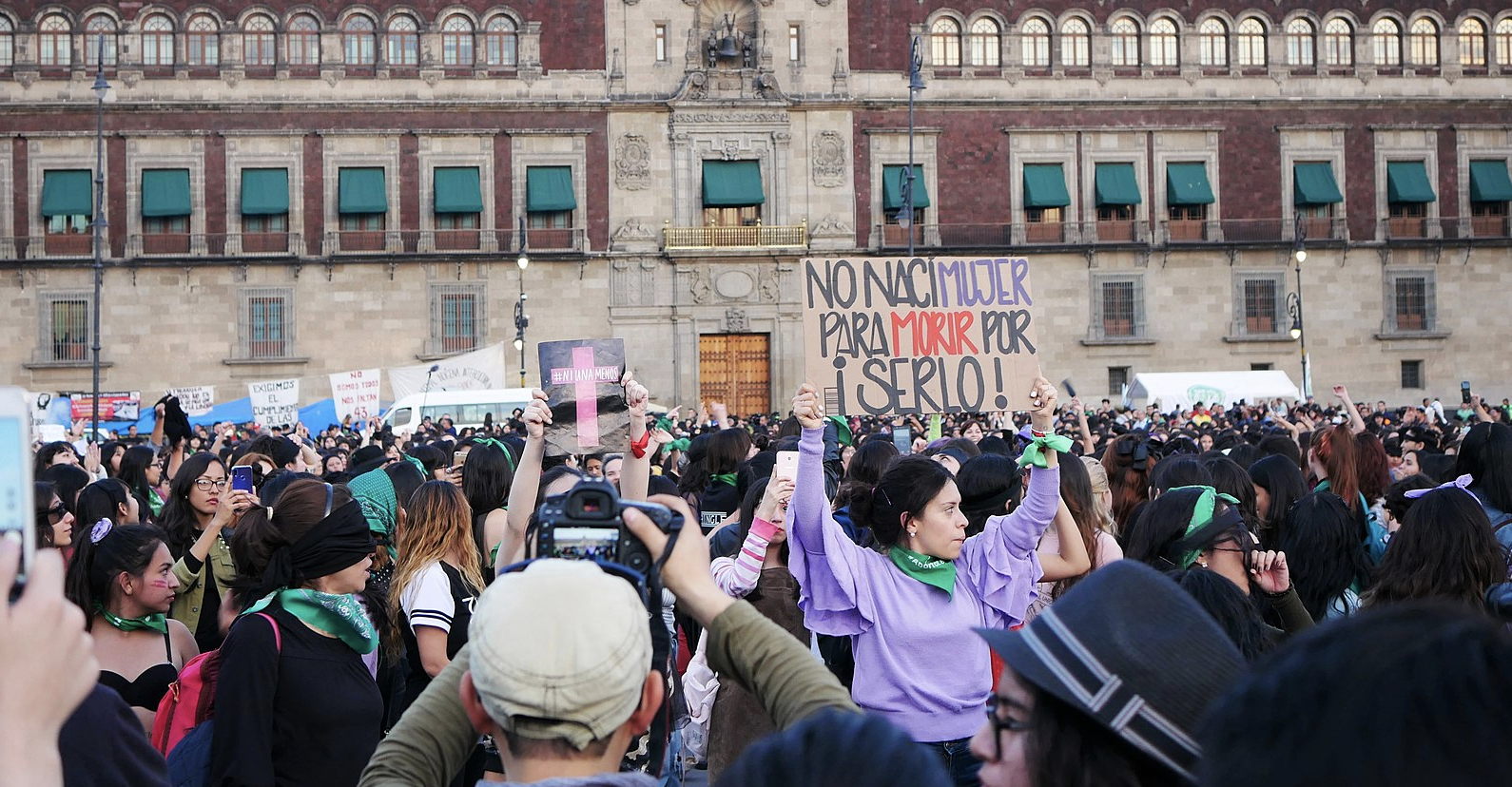 A woman stands in a crown in front of an ornate building, holding a sign that reads (in Spanish) 