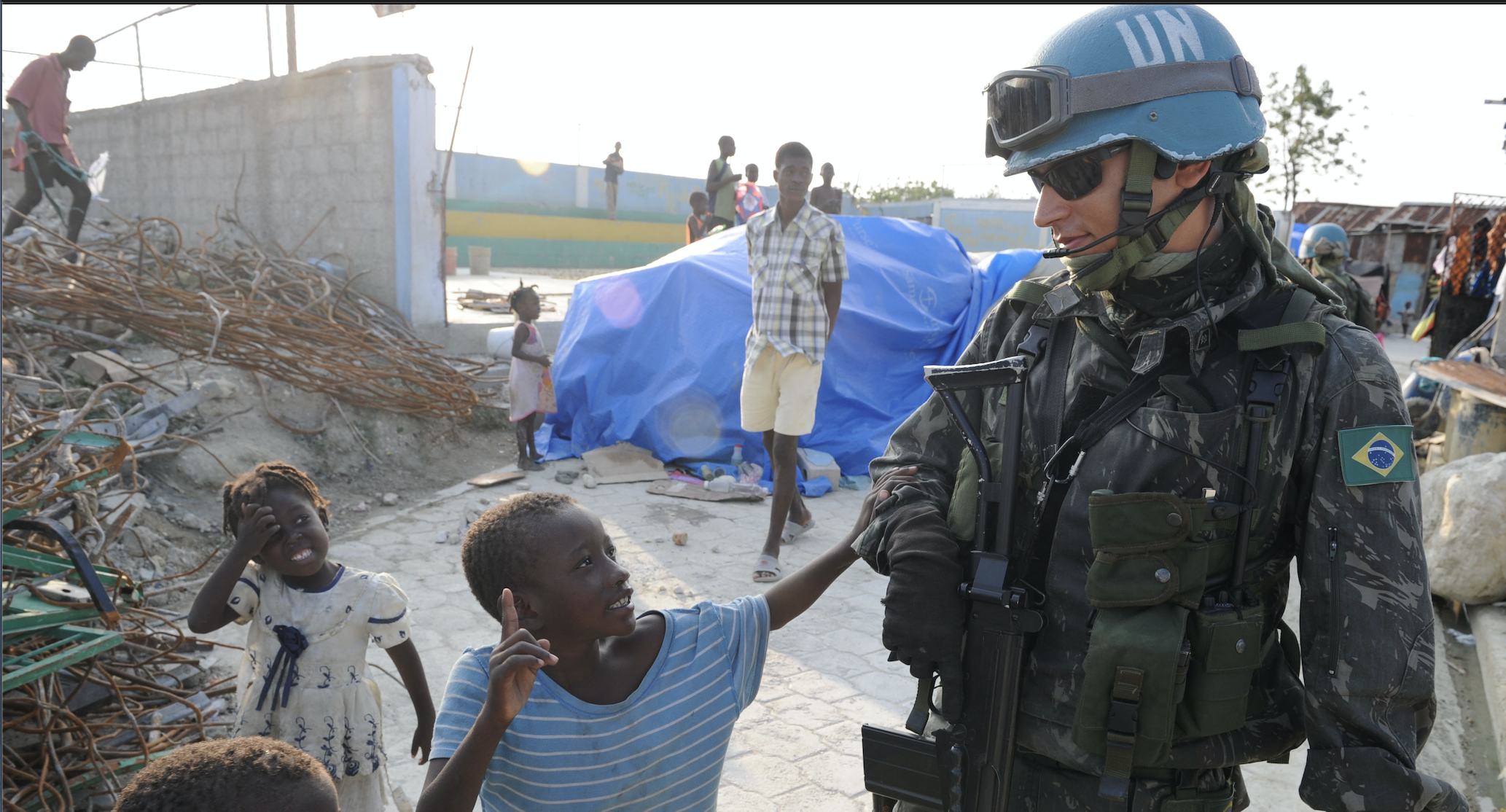 A tall male wearing a UN peacekeeper helmet says hello to some small children in Brazil.
