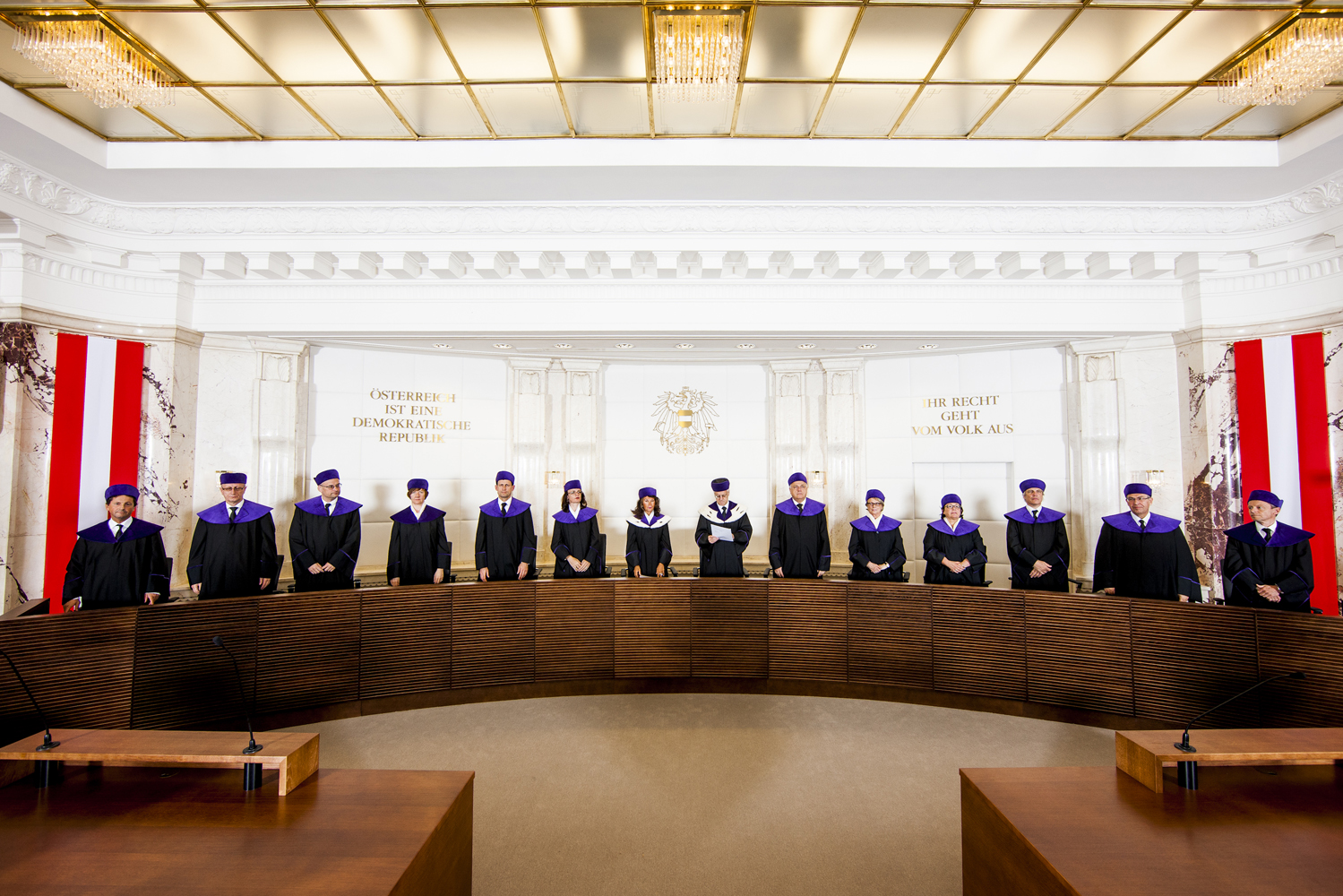 Members of the Austrian constitutional court stand behind a long wooden table flanked by the Austrian flag.
