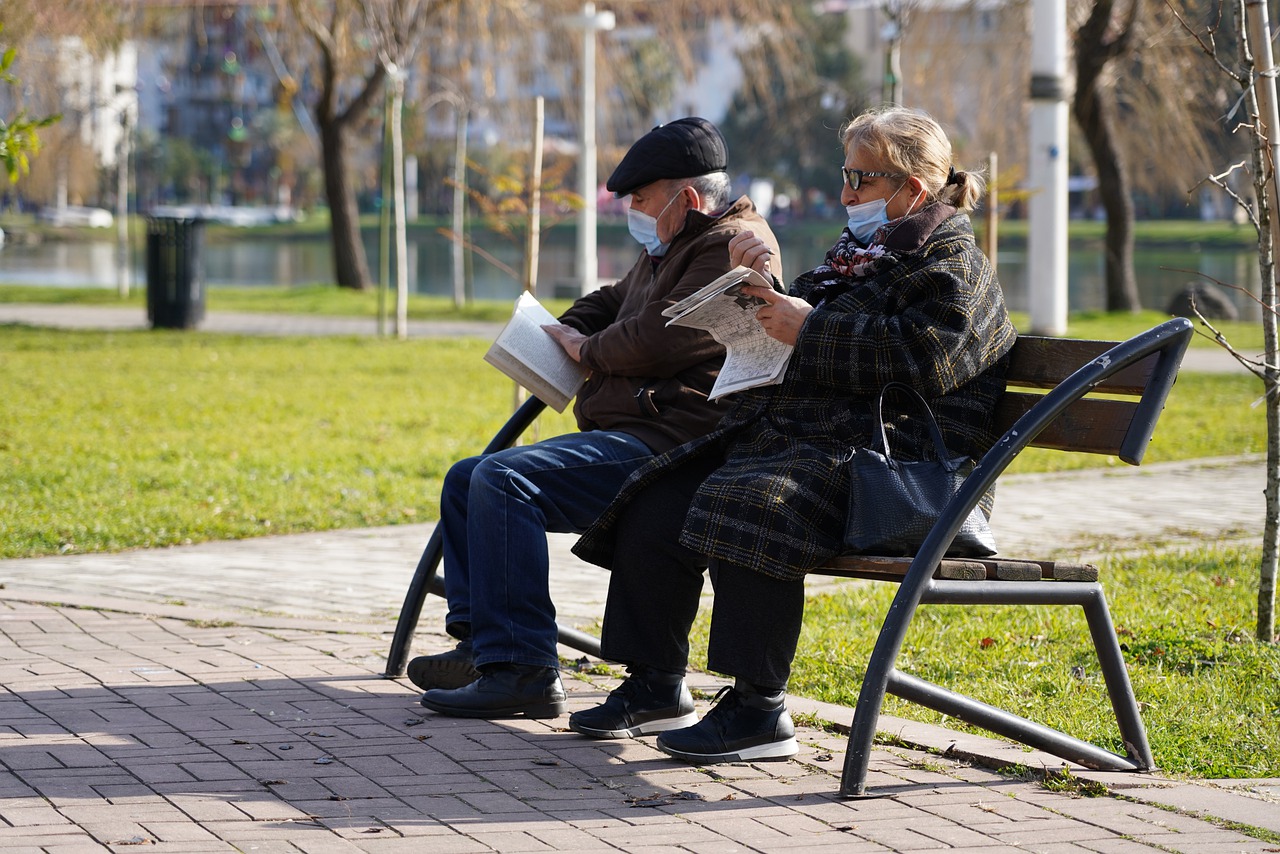 two elderly people with masks on sit on a bench outside