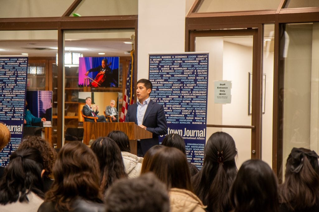 A man speaking to an audience in a formal setting, with banners and framed photos behind him.