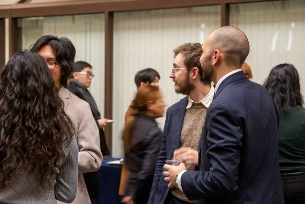 People conversing at a formal event, dressed in business attire, with others mingling in the background.