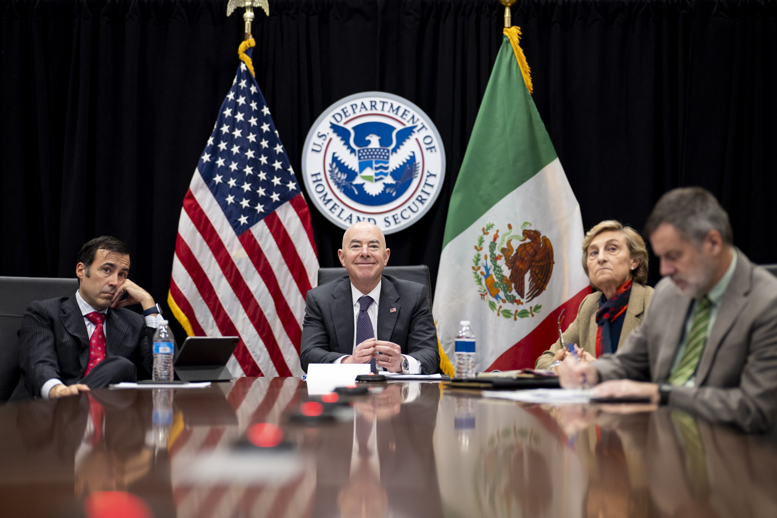 Homeland Secretary seated on a table with Mexico's Secretary of National Defense, General Luis Cresencio Sandoval, and the US Ambassador to Mexico, Ken Salazar. US and Mexico flags can be seen in the background.